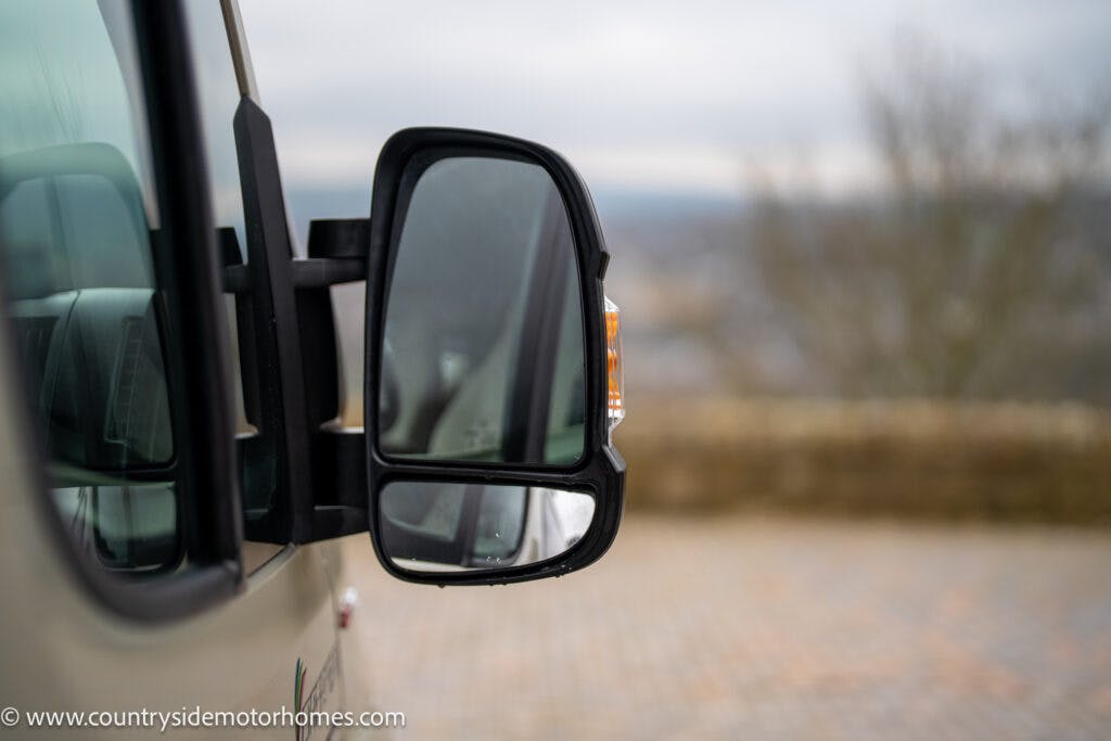 Close-up of the side mirror of a 2020 Rapido Dreamer Select Campervan XL, showing a clear reflection of the surrounding area. The background is blurred, highlighting the mirror. The vehicle is parked on a paved surface with a stone wall and trees visible in the distance.