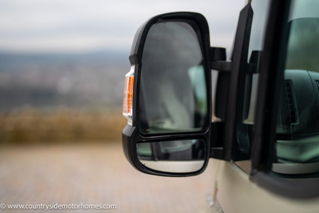 Close-up of a side mirror on a 2020 Rapido Dreamer Select Campervan XL. The reflection shows a blurry outdoor background. Visible at the bottom left is the website URL "www.countrysidemotorhomes.com".