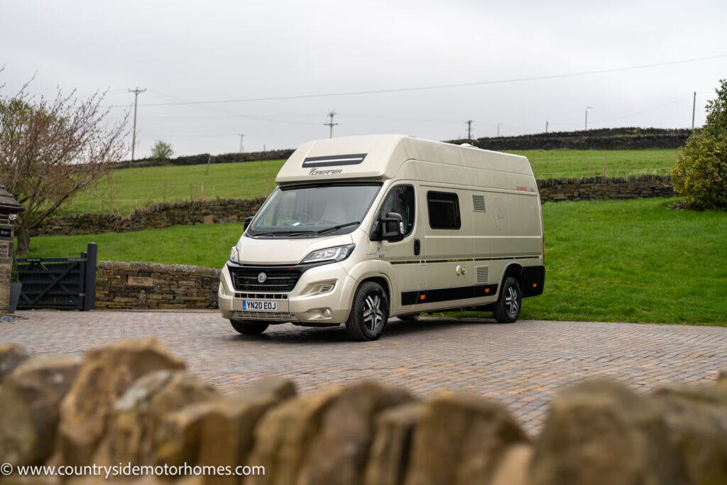 A beige 2020 Rapido Dreamer Select Campervan XL is parked on a paved driveway near a countryside setting with a stone fence. The vehicle features a raised roof and windows along the side. The background includes green grass, a hedge, and an overcast sky.