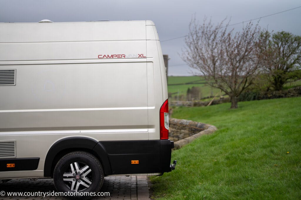 A side view of a parked ivory-colored 2020 Rapido Dreamer Select Campervan XL is visible. It is situated on a grassy area next to a stone wall and pathway. In the background are trees and a misty, overcast sky.