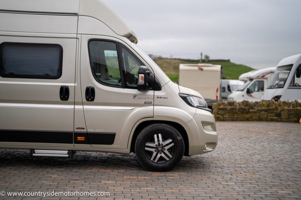 A beige 2020 Rapido Dreamer Select Campervan XL is parked on a cobblestone surface with another white campervan visible in the background. The campervan door is closed, and the surrounding area includes a low stone wall and greenery under a cloudy sky. The URL www.countrysidemotorhomes.com is visible.