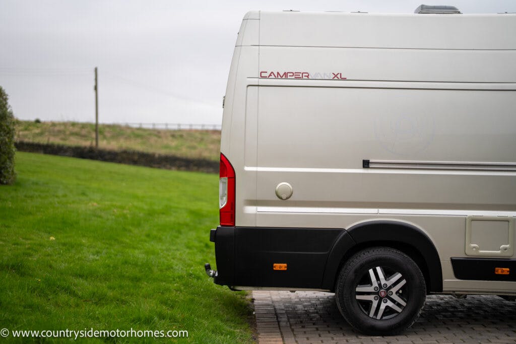 The image shows the rear side of a white 2020 Rapido Dreamer Select Campervan XL parked on a paved area, adjacent to a green lawn. The van displays a red and black logo, and there is a small tow hitch at the back. The background features a cloudy sky and a wooden fence in the distance.