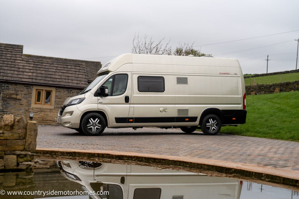 The image features a 2020 Rapido Dreamer Select Campervan XL parked on a paved driveway next to a building with stone walls and a tile roof. The scene is reflected in a calm, shallow body of water in the foreground, against a backdrop of grassy area and overcast sky.