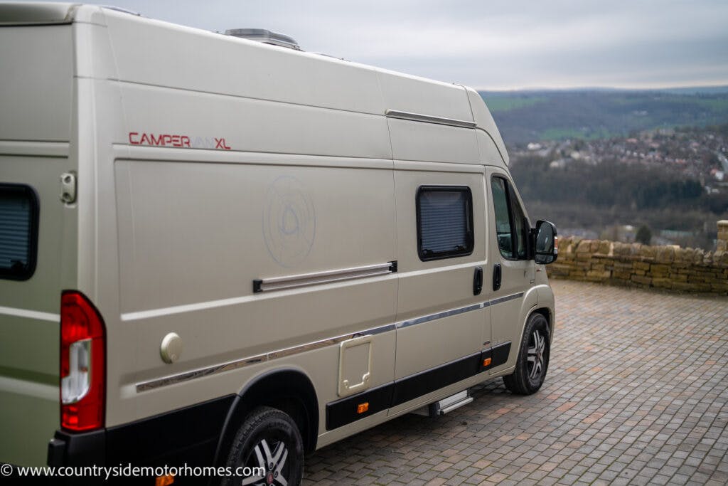 A 2020 Rapido Dreamer Select Campervan XL is parked on a paved area overlooking a rural landscape with distant hills and a cloudy sky. The beige vehicle has a high roof, a logo on the side, and the website “www.countrysidemotorhomes.com” is visible at the bottom left.