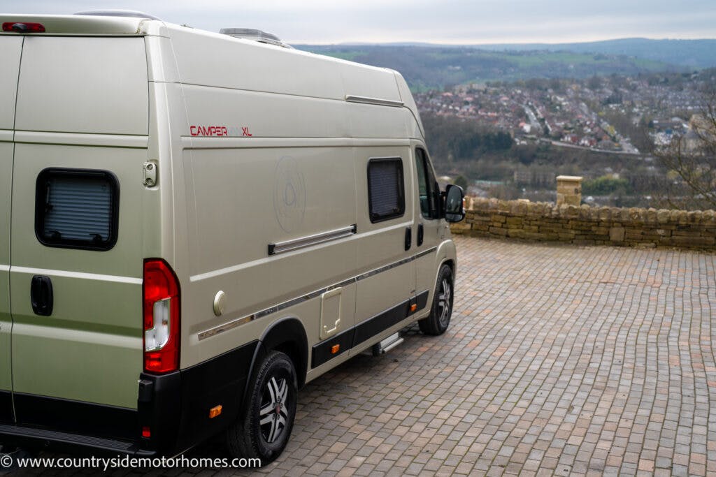 A beige 2020 Rapido Dreamer Select Campervan XL is parked on a paved area with a stone wall and rolling hills in the background. The side of the van features windows, and a website URL is visible at the bottom left corner of the image.