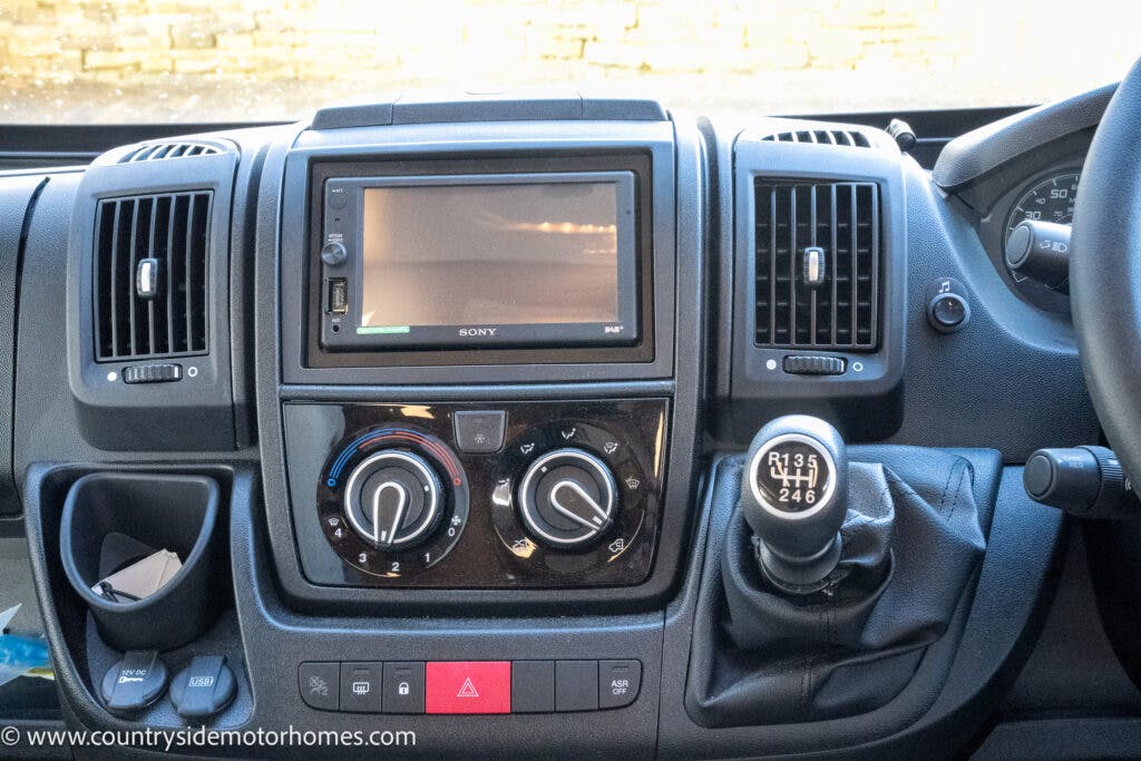 The image shows the dashboard controls of a 2020 Rapido Dreamer Select Campervan XL, featuring a central display screen, air conditioning and heating controls, and a gear shift. Buttons and dials for various functions are also visible, along with part of the steering wheel on the right side.