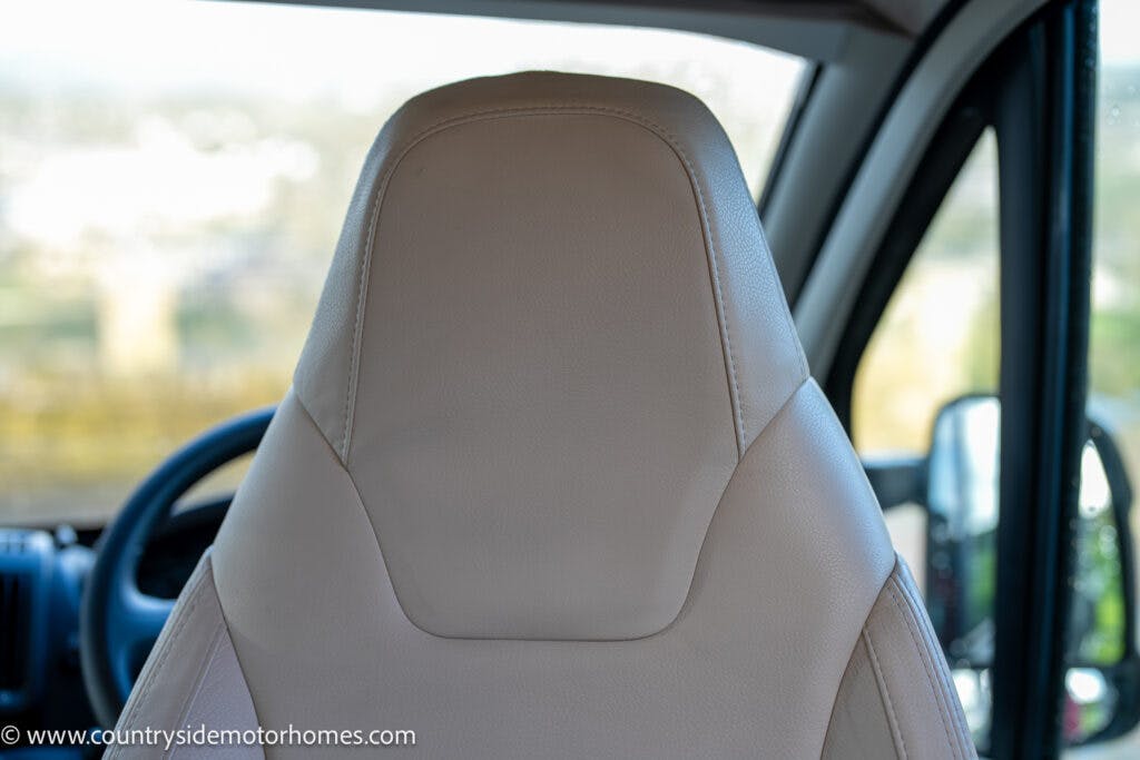 Close-up view of a beige driver's seat inside a 2020 Rapido Dreamer Select Campervan XL. The backside of the seat is visible with the steering wheel and a side mirror slightly blurred in the background. The image has the website www.countrysidemotorhomes.com watermarked at the bottom left corner.
