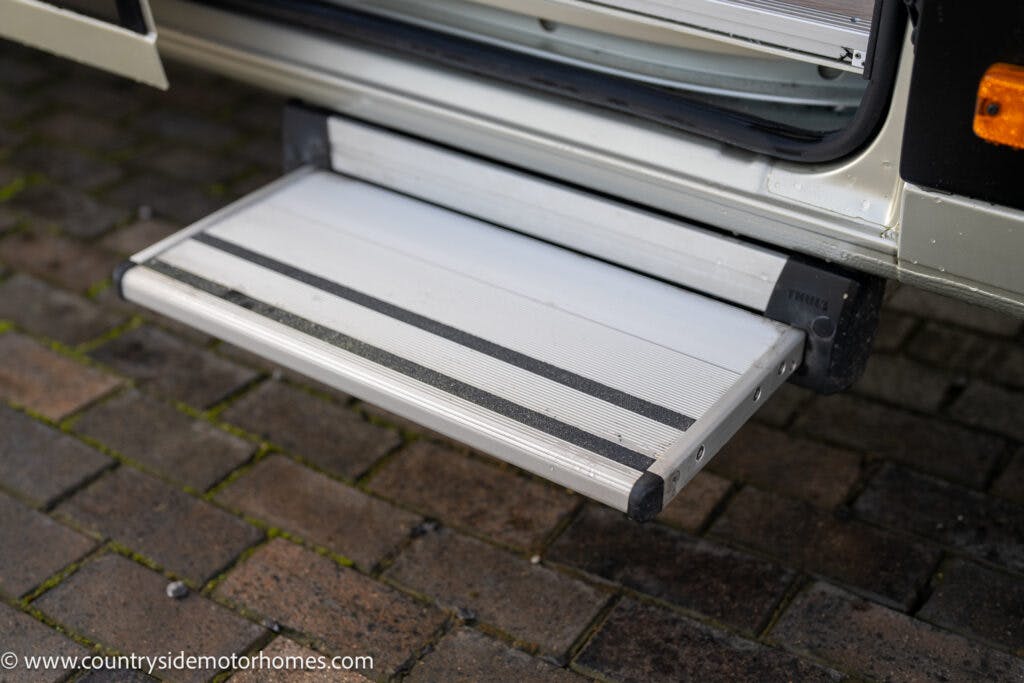 A retractable metal and black rubber step extends from a 2020 Rapido Dreamer Select Campervan XL, positioned over a paved surface. The partially visible door is open, and the step offers a secure foothold for entering or exiting the vehicle. The website URL is visible in the corner.