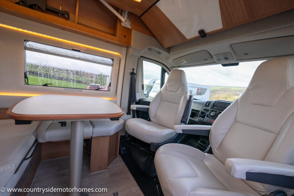 The interior of the 2020 Rapido Dreamer Select Campervan XL features two cream-colored captain's chairs and a matching bench seat around a small dining table. Cabinets above the windows provide storage, while a large window with raindrops offers an inviting view of the outside.