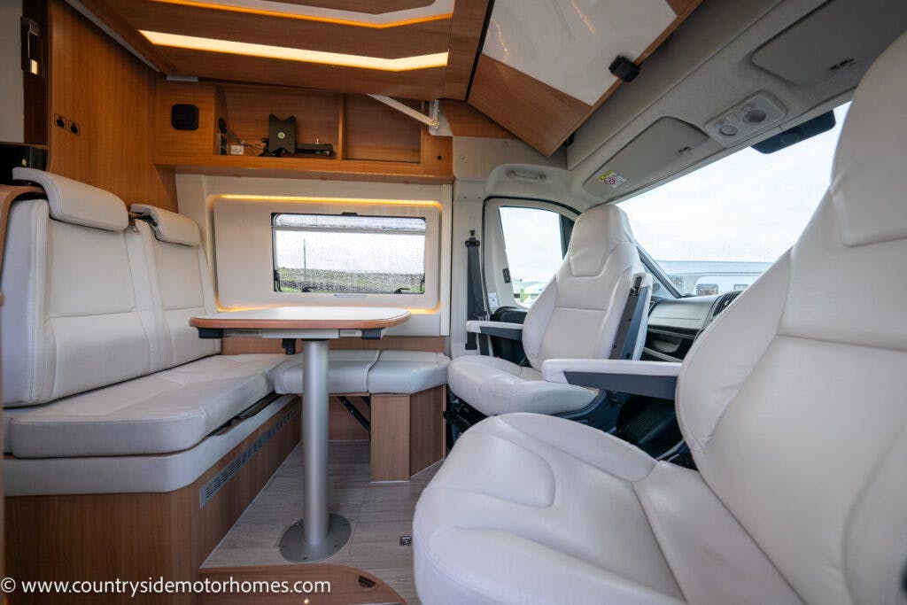 Interior of the 2020 Rapido Dreamer Select Campervan XL with white leather seats. There is a booth-like dining area with a table, a window, and wooden cabinets. The driver's area is visible with the steering wheel and dashboard. The scene is well-lit, showing modern and clean design.