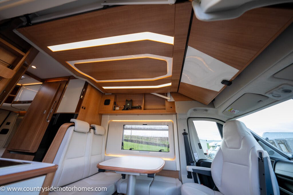 The interior of the 2020 Rapido Dreamer Select Campervan XL features modern wooden cabinetry, a small dining area with a table and two bench seats, white upholstered seating, ambient lighting, and a sleek ceiling with geometric lighting. A large window provides natural light.

