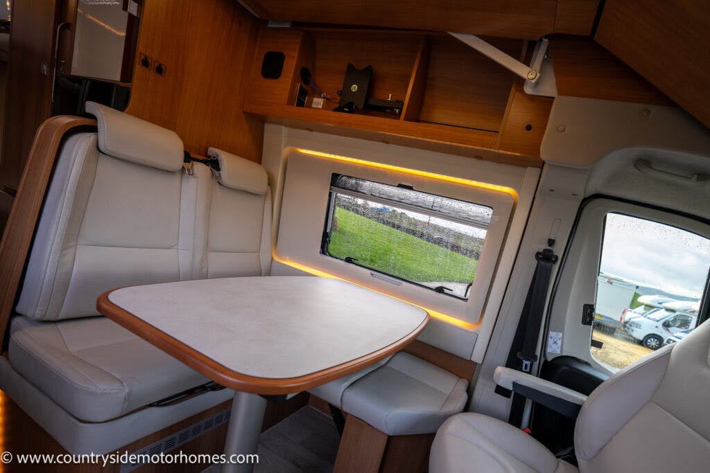 Interior of a 2020 Rapido Dreamer Select Campervan XL featuring a small dining area with a table and cushioned seats. The seating area is next to a window with scenic outdoor views. The space is well-lit with ambient lighting and wooden cabinetry. The vehicle brand is visible on the bottom left.