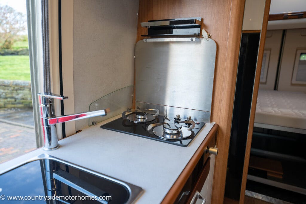 This image shows the kitchen area of a 2020 Rapido Dreamer Select Campervan XL. There is a sink with a modern faucet on the left and a two-burner gas stove with a metal backsplash on the right. The countertop is clean and minimalistic, with the rest of the interior visible in the background.
