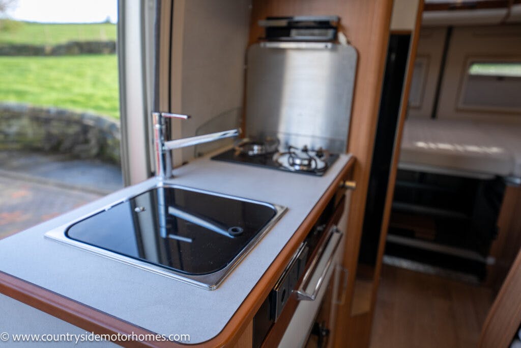 Interior of a 2020 Rapido Dreamer Select Campervan XL showing a compact kitchen area. There is a sink with a modern faucet, a gas stove with two burners, and an overhead range hood. Behind the kitchen area, part of a bed is visible. The URL www.countrysidemotorhomes.com is in the lower-left corner.