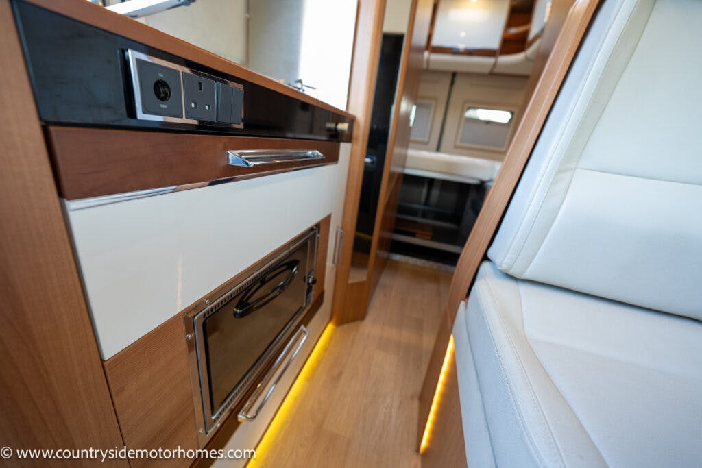 Interior view of the 2020 Rapido Dreamer Select Campervan XL showing a kitchen area with modern cabinetry, an induction cooktop, and a microwave oven below the counter. The seating area features a cushioned bench. Warm ambient lighting is visible near the floor.