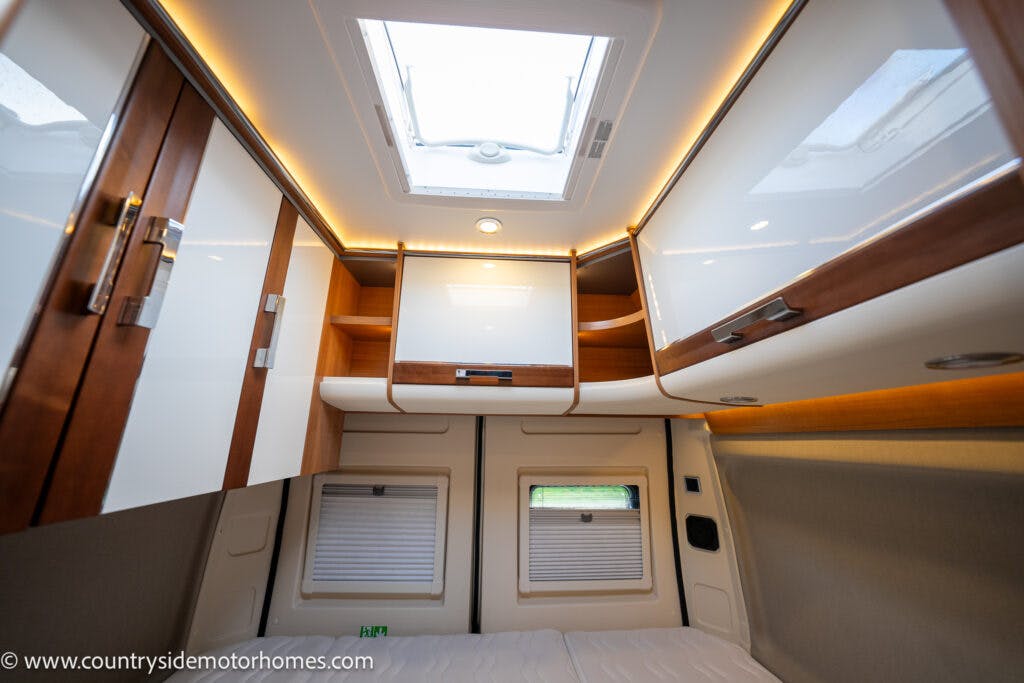 Interior view of the 2020 Rapido Dreamer Select Campervan XL bedroom featuring a double bed with a light beige mattress, white and wooden storage cabinets above the bed, and two small windows with blinds on the far wall. A skylight in the ceiling provides additional natural light.