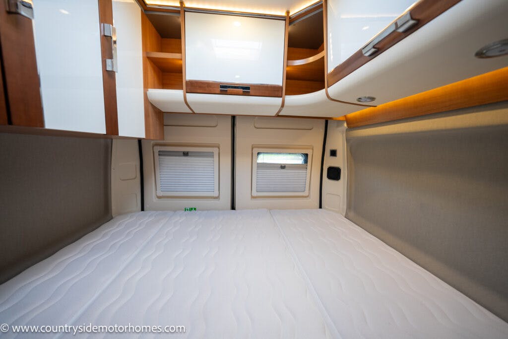 Interior of a 2020 Rapido Dreamer Select Campervan XL bedroom with a neatly made double bed. The room has cabinets with wood and white finishes above and around the bed, with two small windows at the head of the bed. The lighting is soft, and the space appears compact and organized.