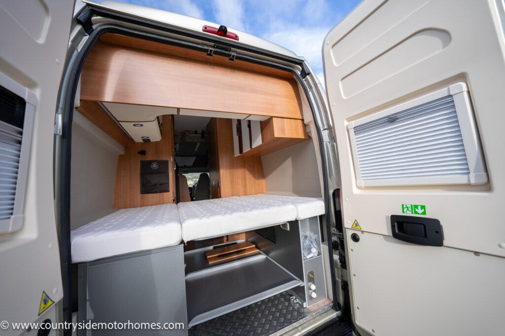Interior view of the rear of the 2020 Rapido Dreamer Select Campervan XL with its doors open, showcasing a bed with white mattress, wooden storage cabinets, and compartments. Lower compartments are open, revealing ample storage space. The van ceiling is visible against a backdrop of the blue sky outside.
