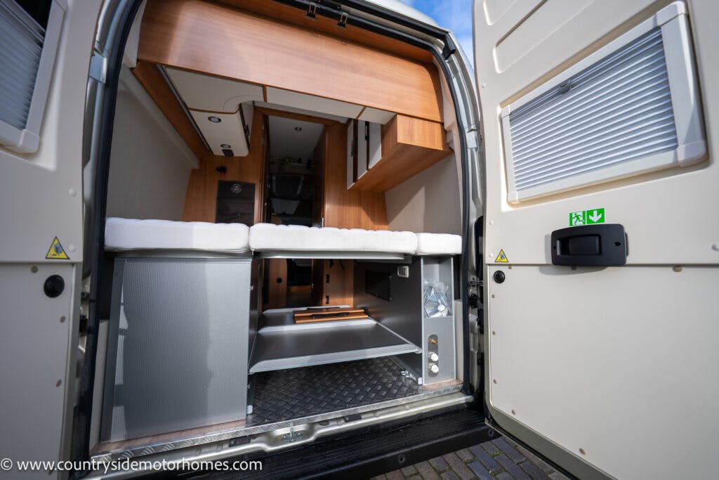 The image shows the open rear doors of a 2020 Rapido Dreamer Select Campervan XL, revealing the interior. The visible design includes wood finishes, storage compartments, a seating area with cushions, and various fixtures. The bottom section has a metallic storage area with a checker plate floor.