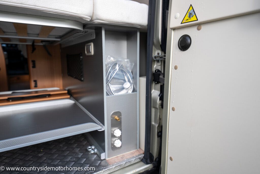 Interior view of a 2020 Rapido Dreamer Select Campervan XL, showing storage compartments. The left side features a gray shelf with a mesh pocket and plastic-wrapped items. The right side has a small cubby with two circular openings. The flooring is textured, and a portion of the bed is visible above.