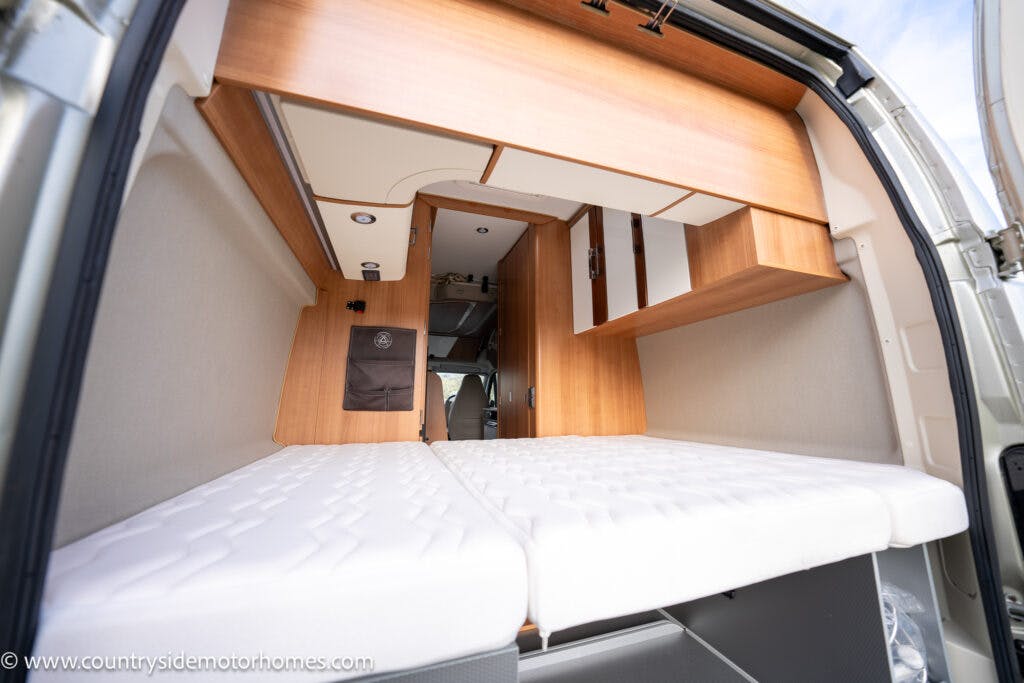 The interior of the 2020 Rapido Dreamer Select Campervan XL boasts wooden cabinetry, featuring a bed with white mattresses and storage compartments above and below. The rear door is open, offering a glimpse into the small, cozy living space.
