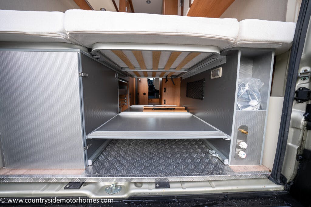 Interior view of the 2020 Rapido Dreamer Select Campervan XL storage compartment accessed through the rear doors. The image displays open cabinets, a sliding shelf, and various storage spaces. The floor has a diamond plate pattern. Wood and metallic finishes are visible.