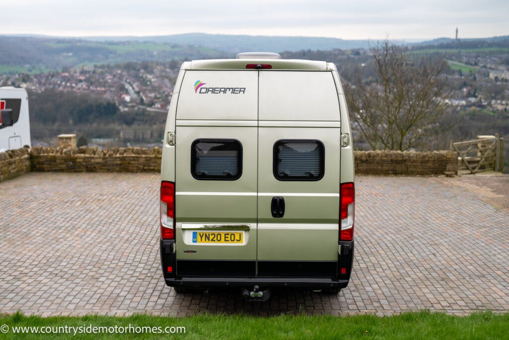 A beige 2020 Rapido Dreamer Select Campervan XL with a license plate reading "YN20 EOJ" is parked on a stone driveway. The campervan features two rear windows with blinds. The background reveals a hilly landscape with scattered buildings. The motorhome has the logo and text "Dreamer" on the back.