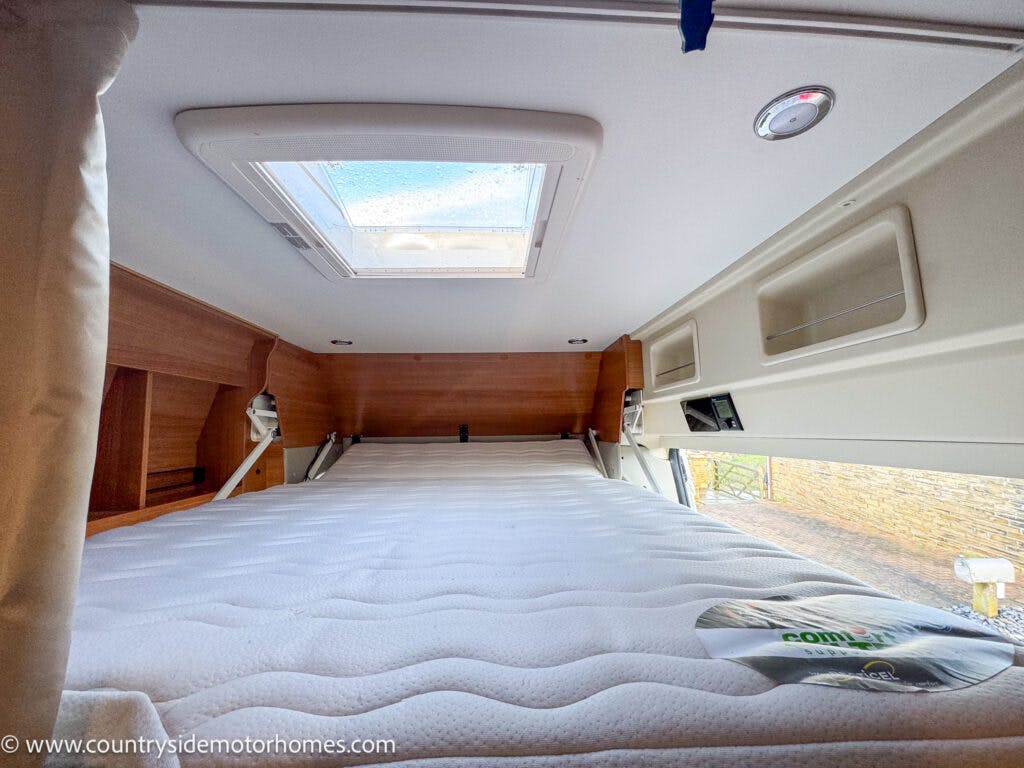 An interior view of a 2020 Rapido Dreamer Select Campervan XL's sleeping area. It features a mattress beneath a skylight window, surrounded by wooden paneling and built-in storage compartments. A label is visible on the mattress, and the website www.countrysidemotorhomes.com is in the bottom left corner.