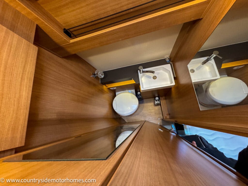 A small bathroom with wooden paneling, inside a 2020 Rapido Dreamer Select Campervan XL, featuring a compact white sink with a chrome faucet, a white toilet, and a mirrored wall. The sink and toilet are adjacent, and there is a partial view of the shower area.