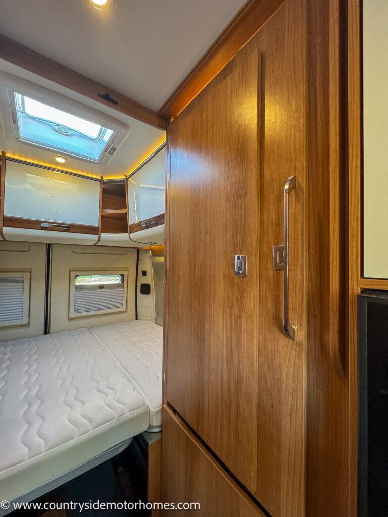 The image shows the interior of a 2020 Rapido Dreamer Select Campervan XL with wooden cabinets, a bed with a white mattress, and a skylight above. The bed is situated against the rear wall next to two small windows. The cabinets above the bed provide additional storage.