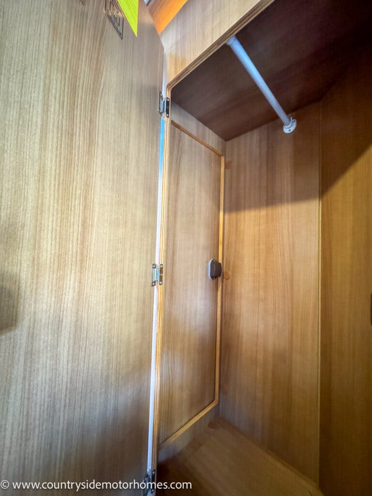 A wooden cupboard with a small door inside stands open, revealing an empty space reminiscent of the storage in a 2020 Rapido Dreamer Select Campervan XL. The interior includes a hanging rod near the top and metal hinges on the doors, marked with a website URL in the bottom left corner.