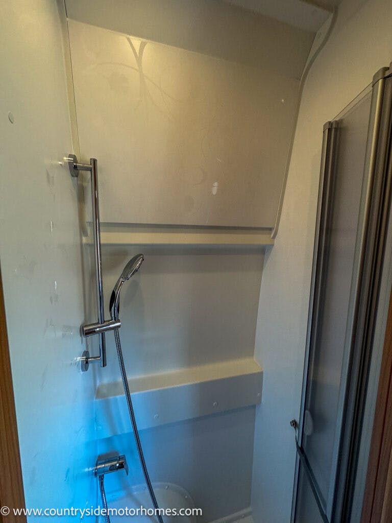The image shows a small shower area in the 2020 Rapido Dreamer Select Campervan XL, featuring a handheld showerhead attached to a vertical bar on the left wall. A translucent folding door is positioned on the right. The website "www.countrysidemotorhomes.com" is visible at the bottom left corner.