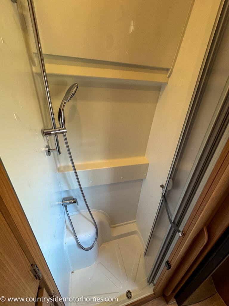 A compact shower area inside the 2020 Rapido Dreamer Select Campervan XL features a handheld showerhead mounted on a vertical bar. The stall has a frosted door on the right, a white interior with a small ledge, and a flexible hose connected to the showerhead. A wooden panel is seen on the left.