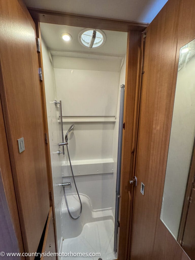 A compact 2020 Rapido Dreamer Select Campervan XL bathroom with a closed wooden door. Inside, there is a small shower area with a handheld showerhead, a shelf, and a circular vent on the ceiling. The interior features white walls and a wooden panel on the side. The mirror is mounted on the door.