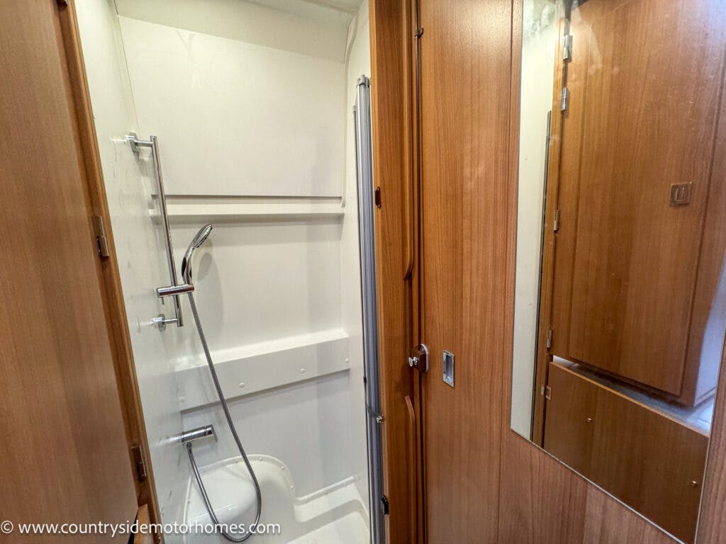 This image shows a compact shower area in the 2020 Rapido Dreamer Select Campervan XL, featuring a white shower stall with a handheld showerhead, a glass door, and wooden cabinetry surrounding the space. There is a mirrored panel adjacent to the shower.
