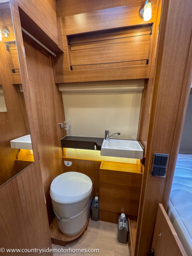The image shows a compact bathroom inside the 2020 Rapido Dreamer Select Campervan XL with wooden finishes. It features a toilet, a sink with a faucet, and a mirror. There are storage compartments above and below the sink. Two bottles are placed on the floor beside the cabinet.