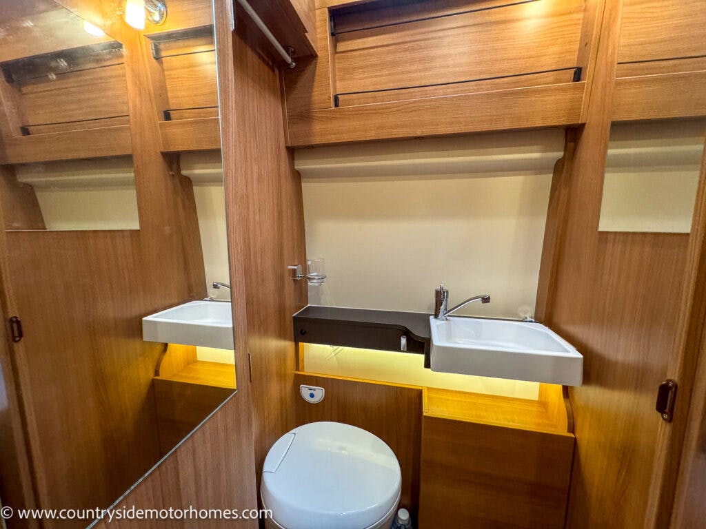 A compact bathroom with wood paneling in the 2020 Rapido Dreamer Select Campervan XL features a small white sink with a single-handle faucet, a glass, and a mirror above it. Below is a white toilet. There are storage cabinets above and below the sink, and the area is well-lit.