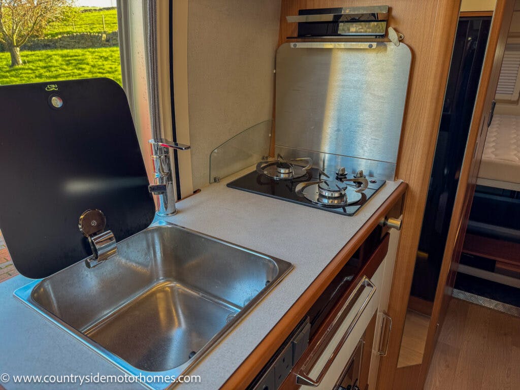 The image showcases a compact kitchen area inside the 2020 Rapido Dreamer Select Campervan XL. It features a stainless steel sink with a faucet on the left and a two-burner gas stove with a metal backsplash on the right. The kitchen has wooden cabinetry and a window overlooking a grassy area outside.
