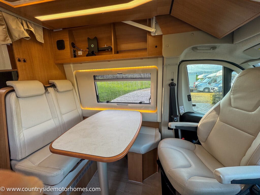 The interior of the 2020 Rapido Dreamer Select Campervan XL features a compact dining area with a table surrounded by padded seats on three sides, wood finishes, and a window with blinds. Cab seats are visible in the foreground. A website URL is noted at the bottom left corner.