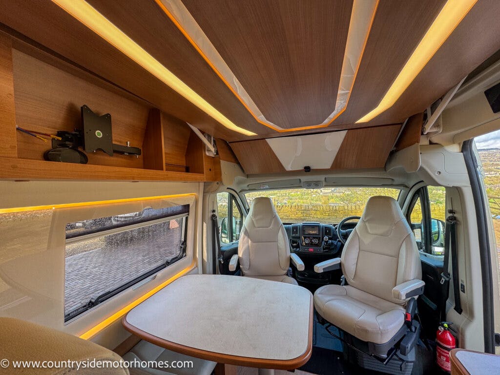 Interior view of the 2020 Rapido Dreamer Select Campervan XL showing a small dining area with two swiveling captain chairs and a table. Overhead cabinets provide storage, and the ceiling features wooden panels with integrated lighting. The driver's cab is visible in the background.