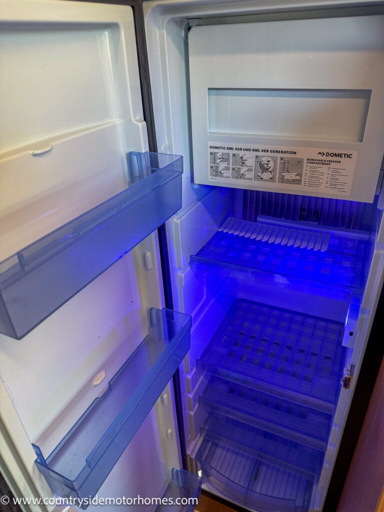Interior view of an empty refrigerator with a closed freezer compartment at the top. The shelves and door compartments are illuminated with blue lighting. A sign inside the refrigerator references the Dometic brand and other information specific to the 2020 Rapido Dreamer Select Campervan XL.
