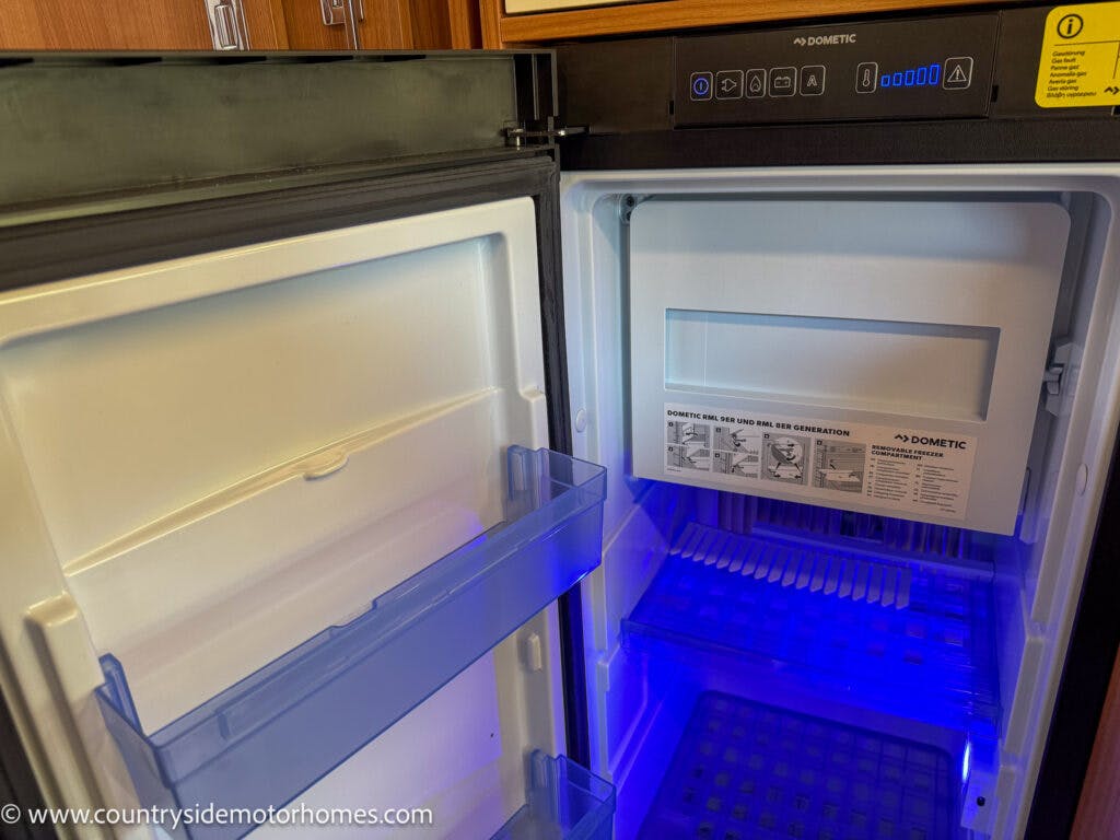 An open Dometic refrigerator inside a wooden cabinet in the 2020 Rapido Dreamer Select Campervan XL. The fridge door features two transparent shelves, while the interior has a cool blue light, an ice box with an instruction sticker, and a grid shelf at the bottom. A website URL is visible at the bottom left.