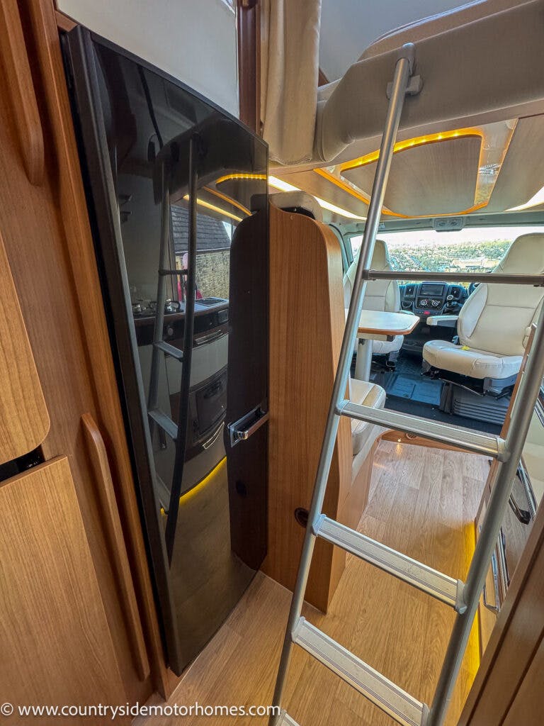 The interior of a 2020 Rapido Dreamer Select Campervan XL features a wooden floor, white leather seats in the driver's area, a black refrigerator, and wooden cabinets. A metal ladder leads up to an overhead bunk. The website www.countrysidemotorhomes.com is visible at the bottom left.