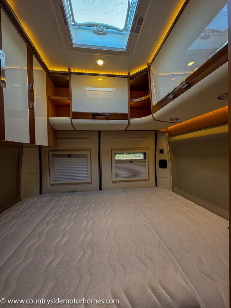 The image shows the interior of a 2020 Rapido Dreamer Select Campervan XL bedroom with storage cabinets, a skylight, and a large bed. The walls and ceiling feature wooden and white glossy finishes. The closed cabinets and natural light from the skylight create an organized and cozy atmosphere.