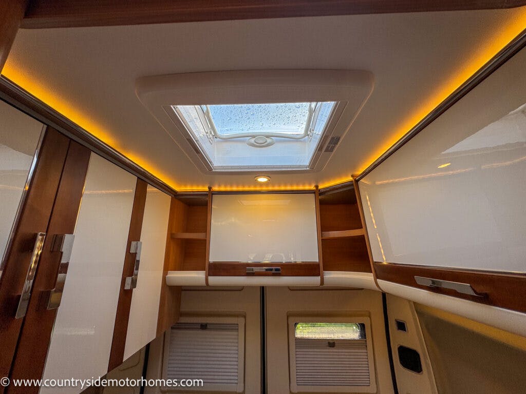         Interior view of a 2020 Rapido Dreamer Select Campervan XL featuring a skylight, surrounded by cabinets with white doors and wooden trim. Warm ambient lighting lines the upper edge where the ceiling meets the walls. The website www.countrysidemotorhomes.com is visible.