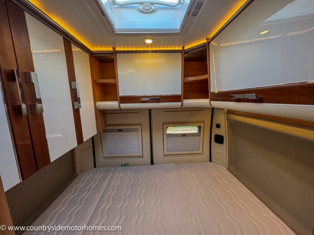 The image shows the interior of a 2020 Rapido Dreamer Select Campervan XL bedroom featuring a fitted mattress, overhead storage cabinets with glossy white doors, wooden accents, and a large skylight above. The walls have beige padding, and there are two small windows at the head of the bed.