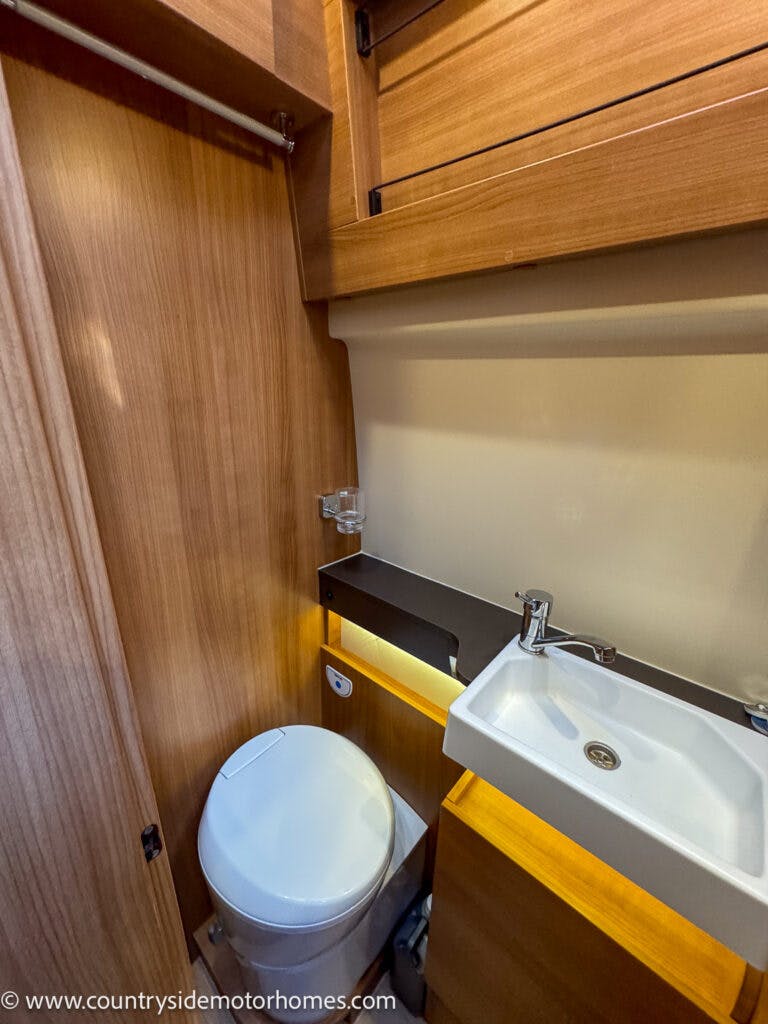 The image shows a compact bathroom inside the 2020 Rapido Dreamer Select Campervan XL. It features a white round toilet on the left and a rectangular white sink with a modern faucet on the right. The walls and cabinetry are made of light wood, and there is a small shelf above the sink.