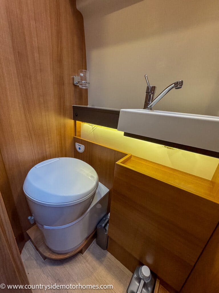 The compact bathroom inside the 2020 Rapido Dreamer Select Campervan XL features a white plastic toilet and a small white sink with a chrome faucet. The walls and cabinetry are wooden, and a strip of lighting below the sink provides illumination. A toothbrush holder is mounted on the wall.