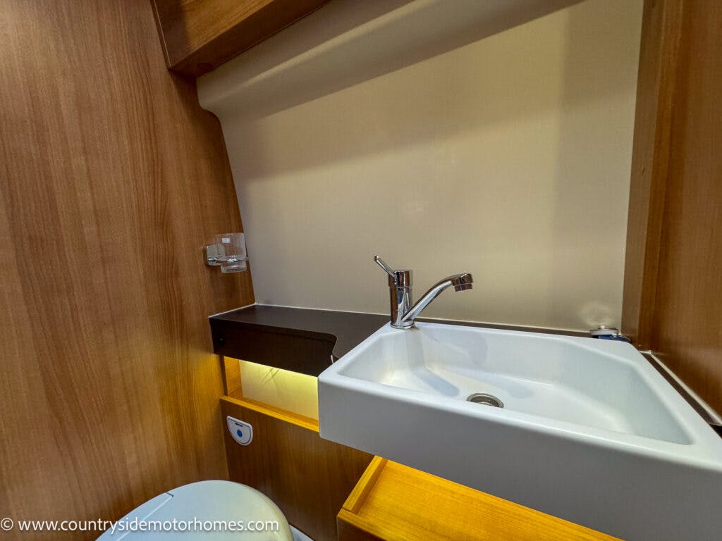 This image shows a compact bathroom sink area inside a 2020 Rapido Dreamer Select Campervan XL. The white sink has a chrome faucet and is set against a wooden wall. Beneath the sink is a lit wooden shelf, and to the right is an acrylic soap dish. The toilet is partially visible on the left.