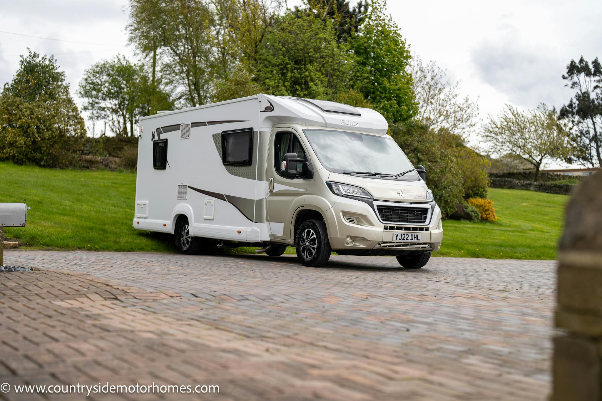 A 2022 Elddis Autoquest 150 Lombardi motorhome is parked on a paved area near a grassy lawn with trees in the background. The cream and tan vehicle features a side door and several windows. The scene appears overcast with a mix of trees and a stone wall in the distance.